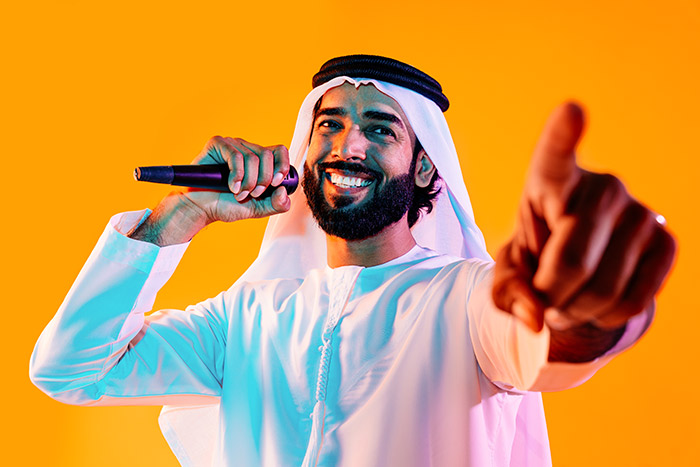 Smiling man in a white kandura and ghutra holding a microphone against an orange background
